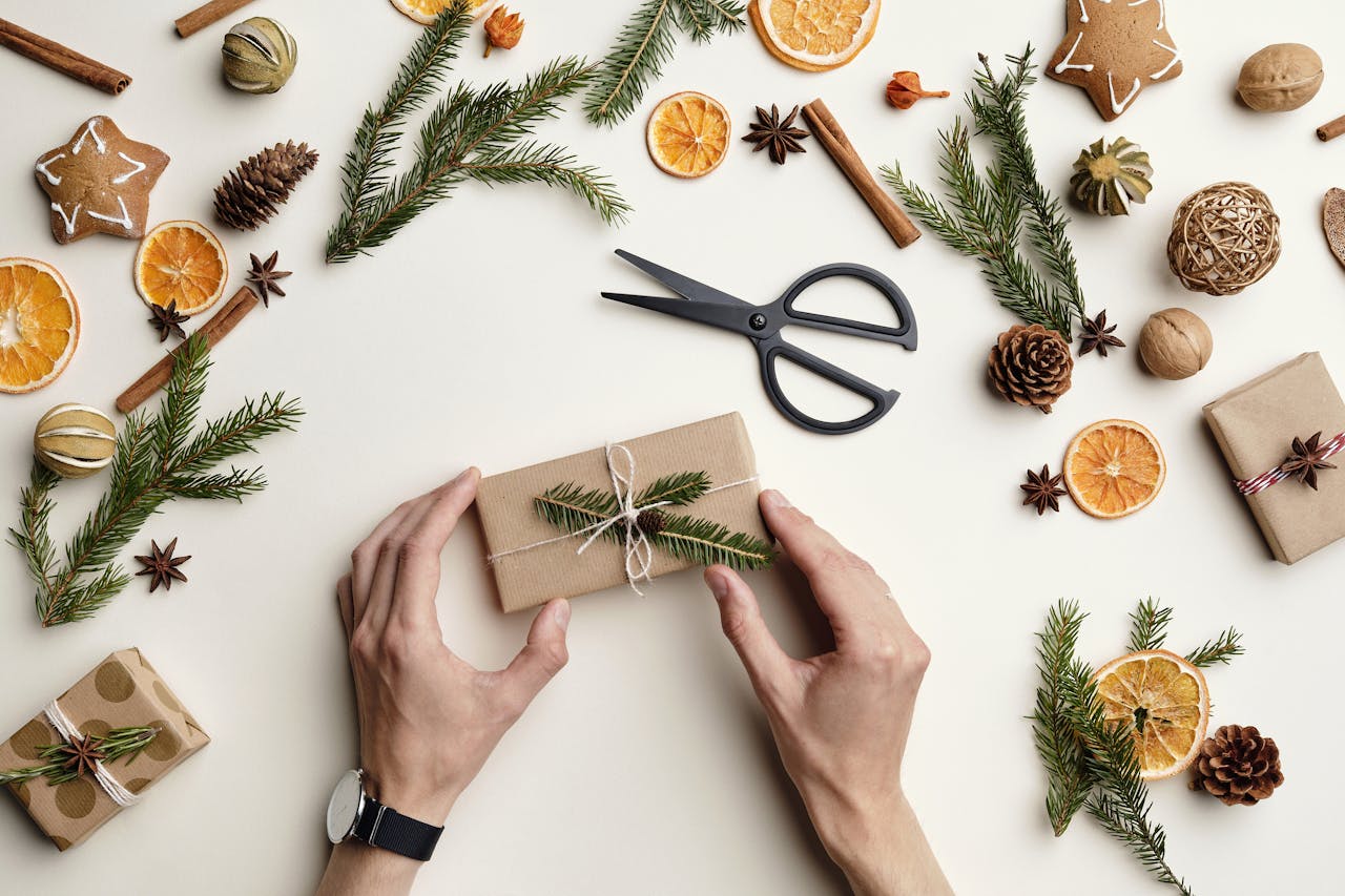 Hands wrapping a Christmas gift with natural decor on a flat white background.